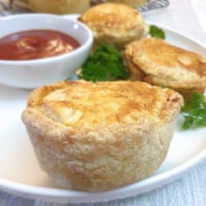 Side on view of a beef, onion and mushroom hand pie on a plate with a dish of tomato sauce in the background.