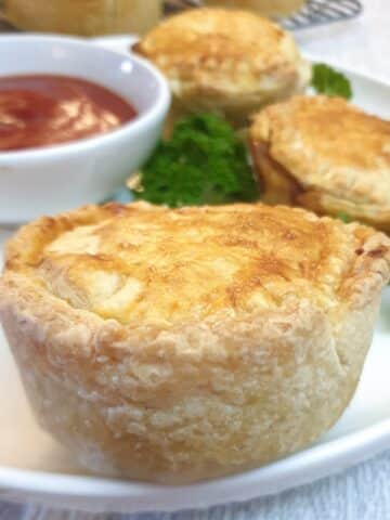 Side on view of a beef, onion and mushroom hand pie on a plate with a dish of tomato sauce in the background.