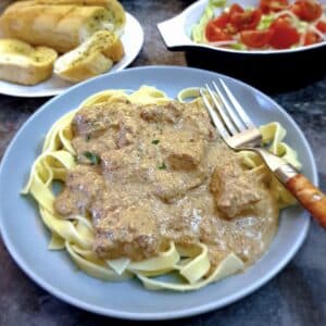 A blue plate of chicken with creamy garlic and mushroom sauce with tomatoes and garlic bread in the background.