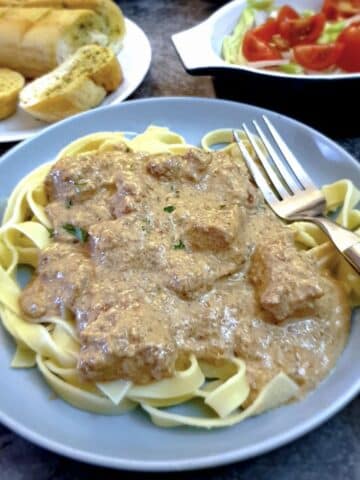 A blue plate of chicken with creamy garlic and mushroom sauce with tomatoes and garlic bread in the background.