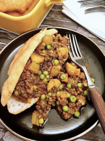 A vetkoek which has been split open and filled with curried mince on a brown plate with a fork.