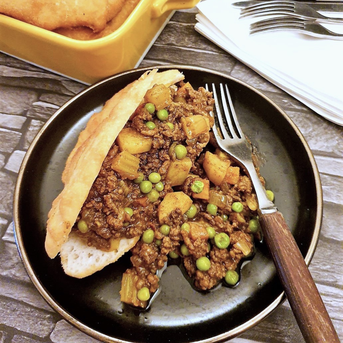 A vetkoek which has been split open and filled with curried mince on a brown plate with a fork.