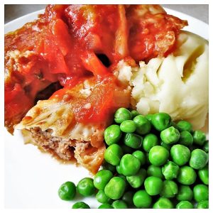 A plate of stuffed cabbage rolls with mashed potatoes and peas.