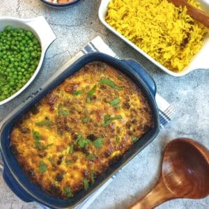 A blue serving dish of bobotie next to a bowl of yellow rice and a bowl of peas.