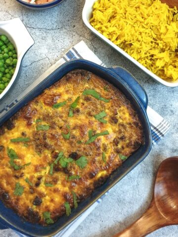 A blue serving dish of bobotie next to a bowl of yellow rice and a bowl of peas.