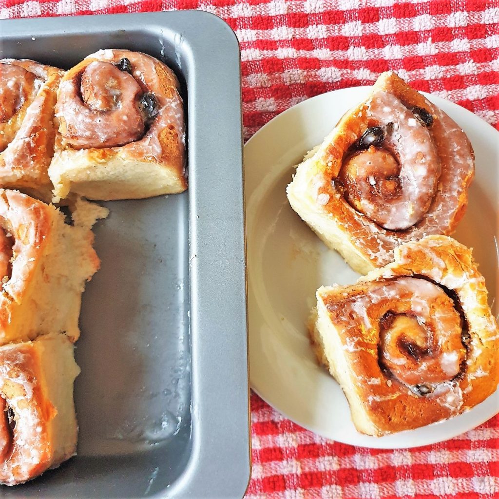 Two chelsea buns on a white plate on a red and white checked cloth.