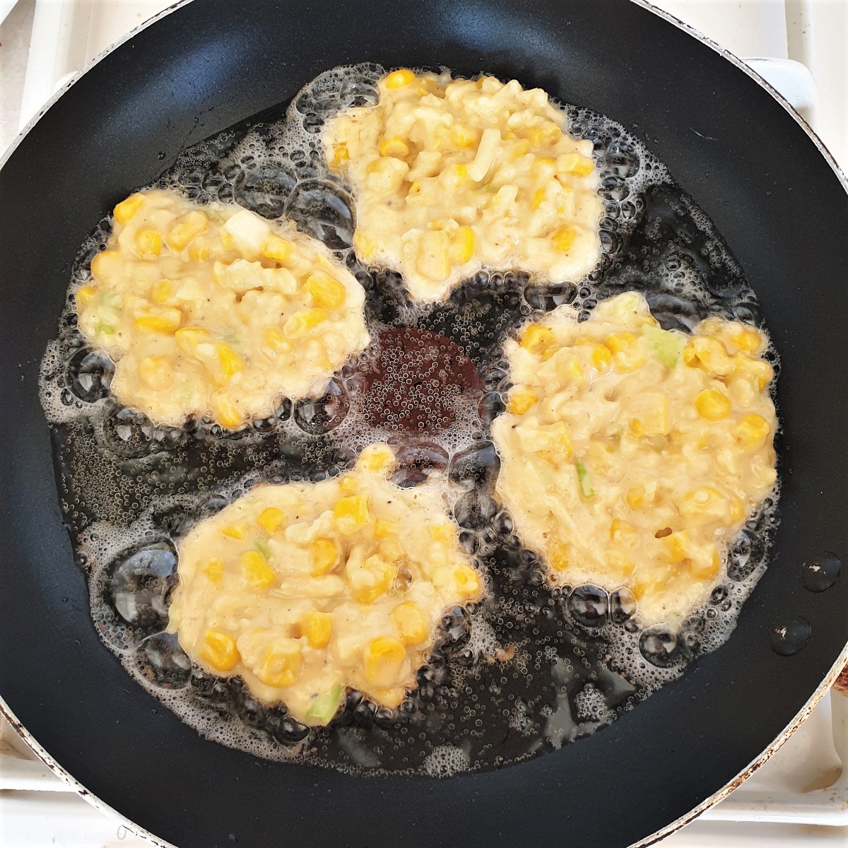 Four sweetcorn fritters frying in a frying pan.