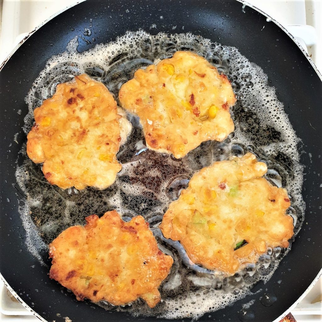 Four sweetcorn fritters browning in a frying pan.