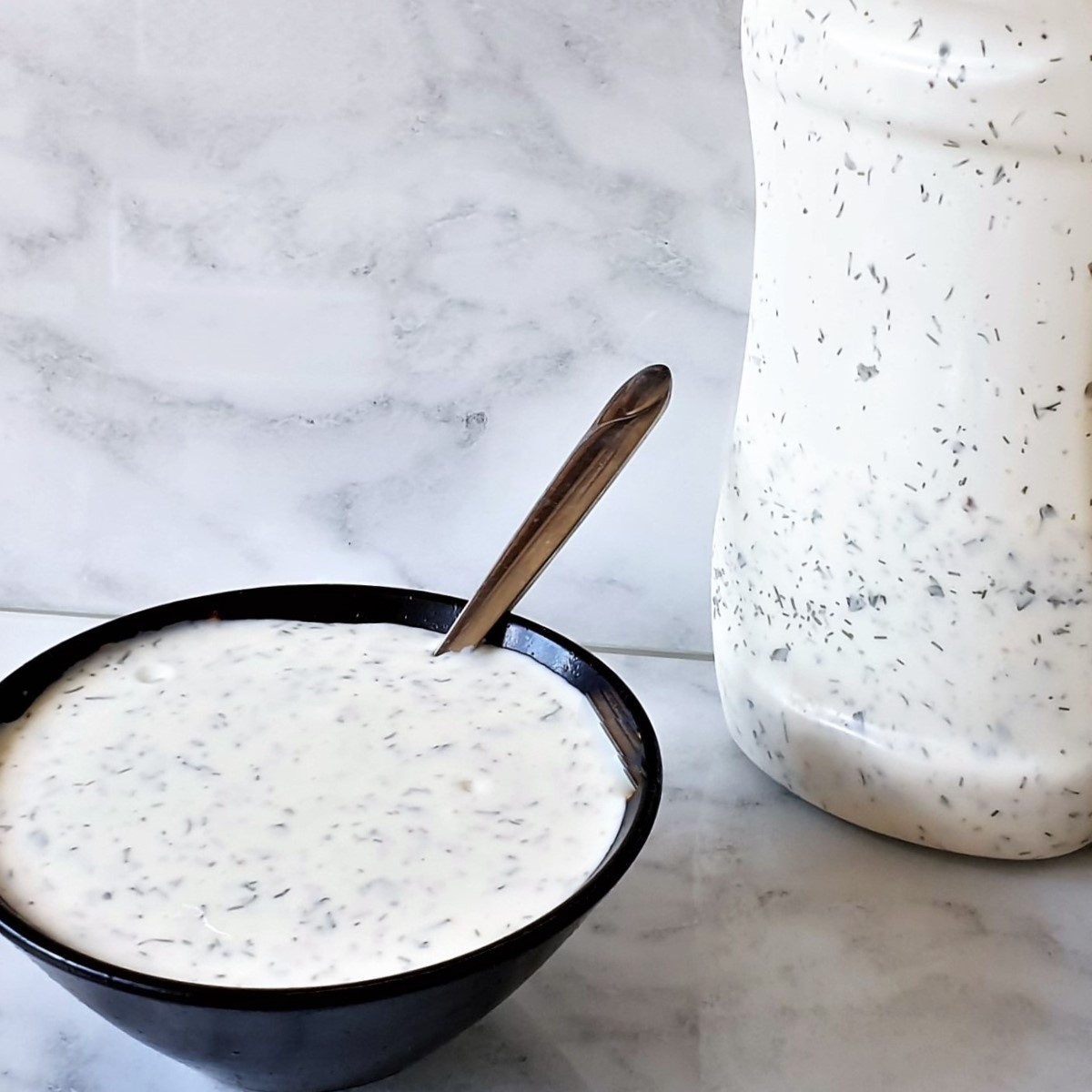 A bowl of homemade ranch dressing next to a bottle of dressing.