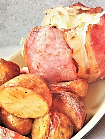 A whole head of cabbage wrapped in bacon on a dish with roast potatoes.
