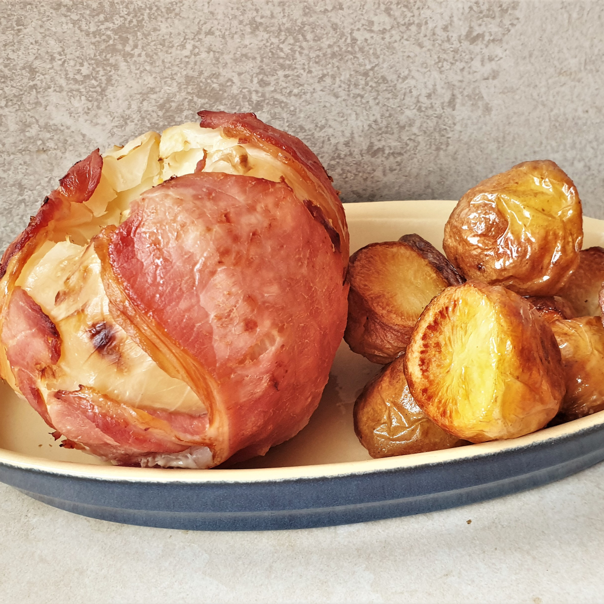 A baked head of bacon wrapped cabbage in a dish with roast potatoes.