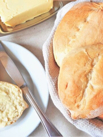 Two Portuguese rolls on a plate next to a roll that has been split open and smeared with butter.