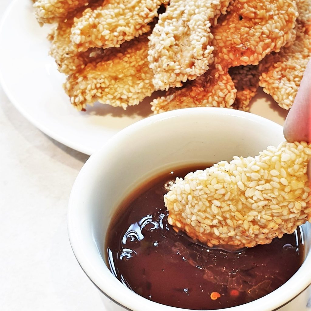 A piece of sesame coated chicken being dipped in a bowl of sauce.
