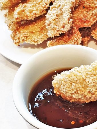 A piece of sesame coated chicken being dipped in a bowl of sauce.