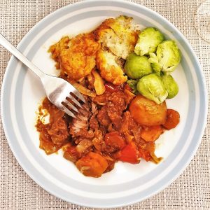 A plate of beef casserole with suet dumplings, showing a fork cutting into the beef, with fluffy dumplings and the first brussels sprouts of the season.