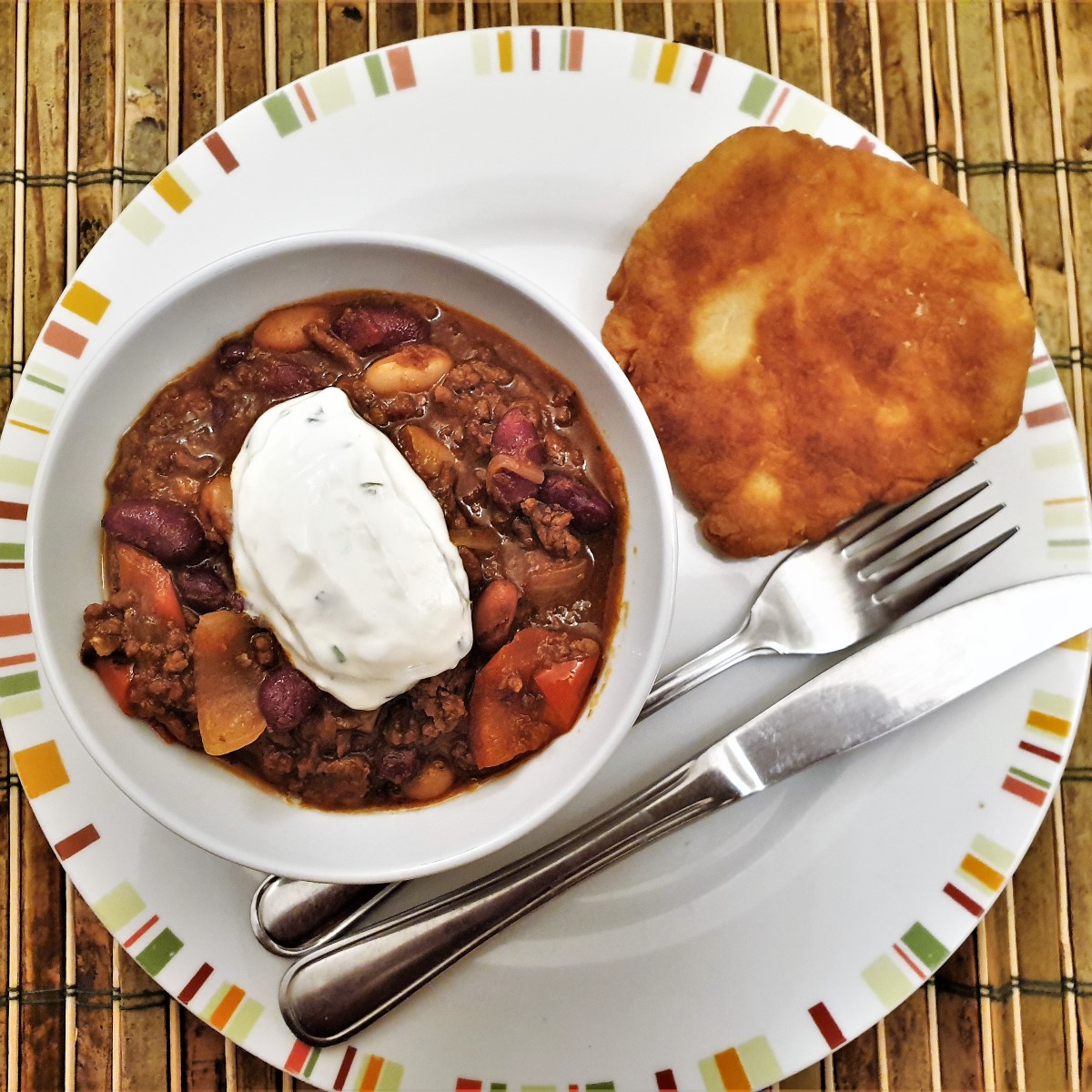 A bowl of chillli con carne topped with sour cream, alongside a homemade vetkoek (or fried dough ball).