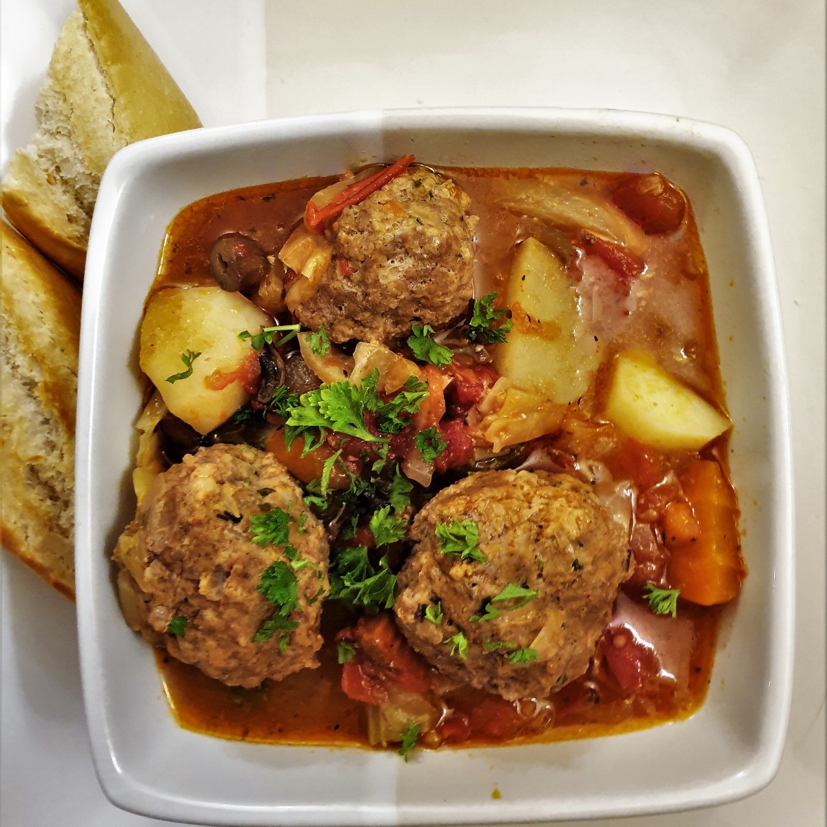 A dish of meatball soup with vegetables and tomatoes and custy bread.