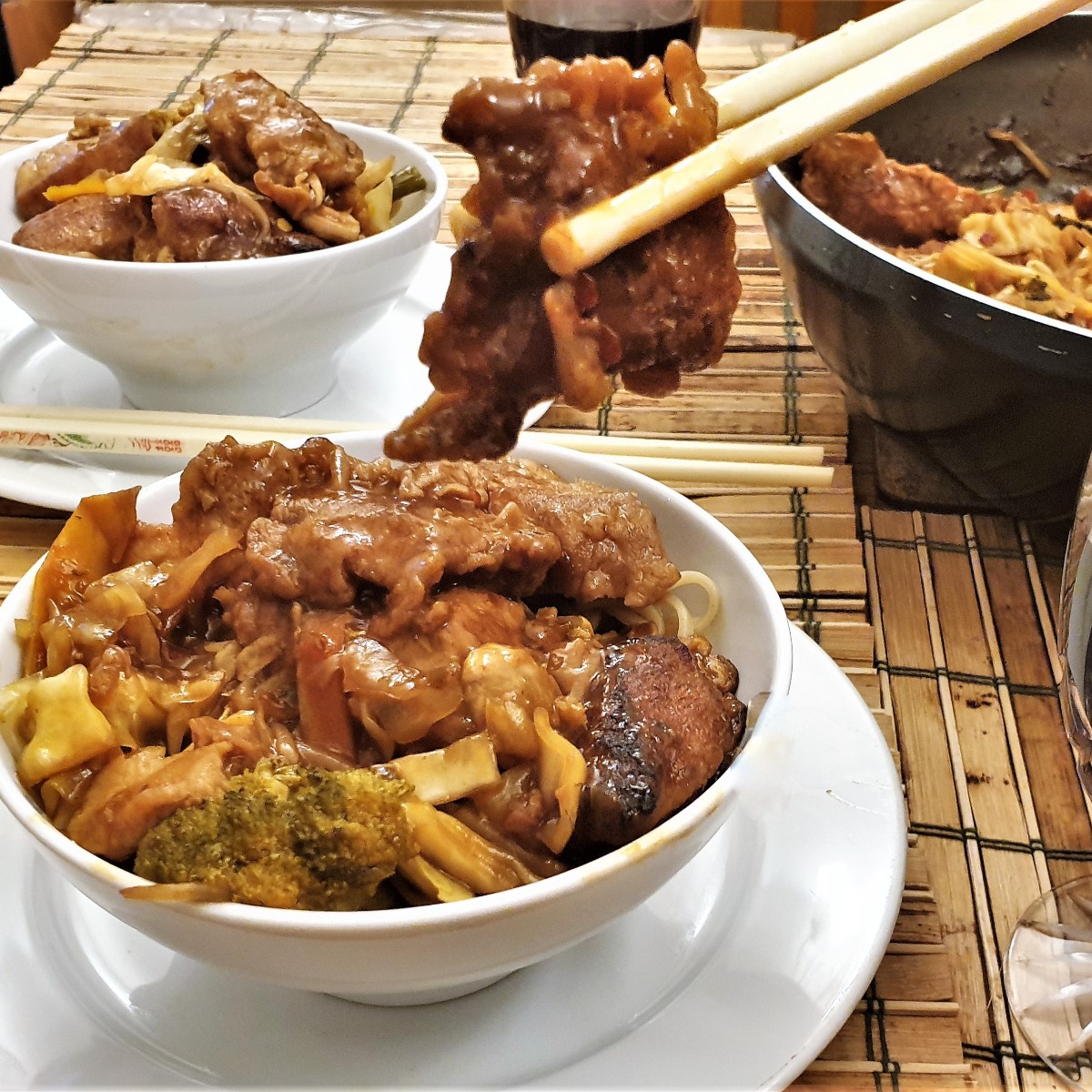 A piece of chicken held with chopsticks above a bowl of szechuan chicken.