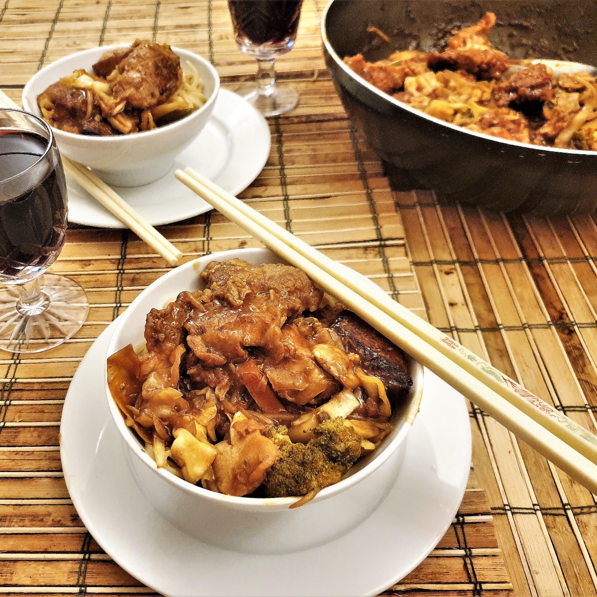 A bowl of szechual chicken with chopsticks in the foreground with a wok of chicken in the background