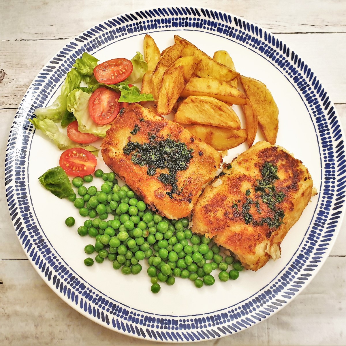 Overhead shot of fried fish fillets on a plate with chips, peas and tomato salad.