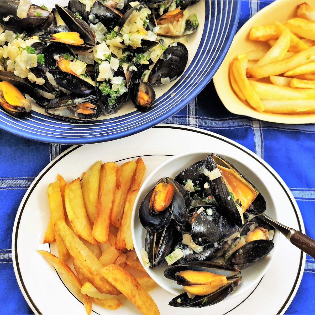 A dish of open mussels on a plate with french fries alongside a serving dish filled with cooked mussels.