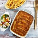 A dish of sausage and bean casserole on a table alongside dish of crispy french fries and a green salad.