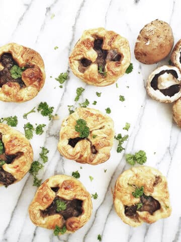 Overhead shot of 6 mini mushroom puff pastry pies. There are a few whole mushrooms scattered between the pies.