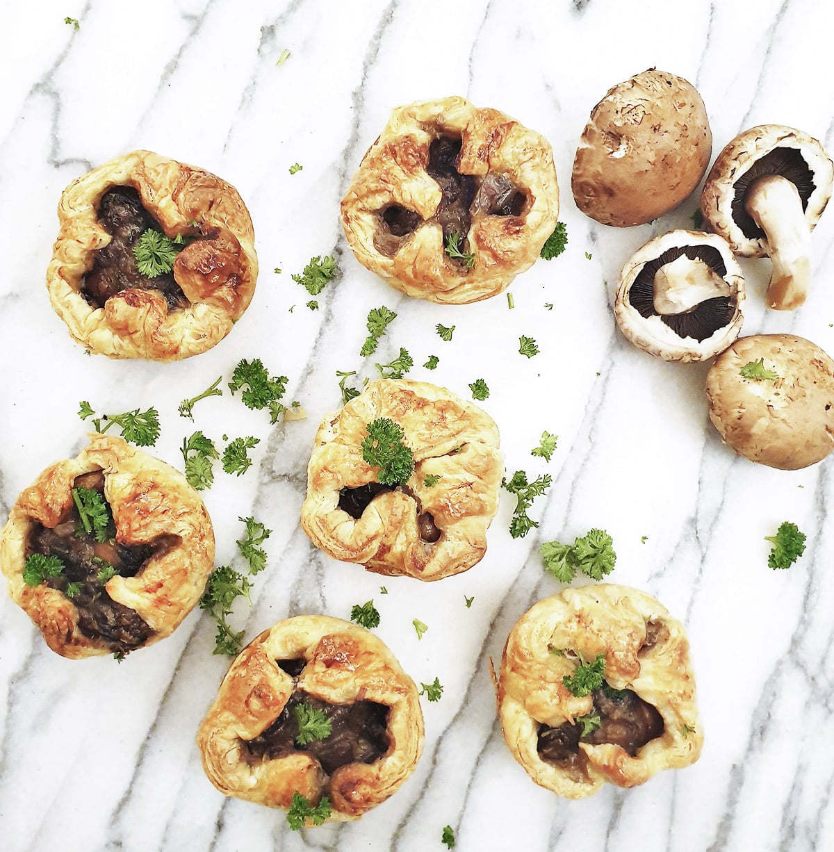 Overhead shot of 6 mini mushroom puff pastry pies. There are a few whole mushrooms scattered between the pies.