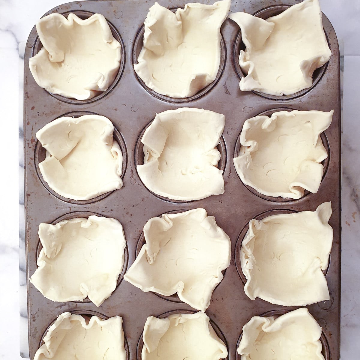 12 squares of puff pastry arranged in the holes of a muffin pan.