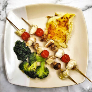 Two monkfish kebabs on a plate with vegetables and potatoes.