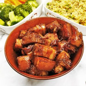 Close up of a dish of sticky pork belly bites with rice and vegetables in the background.