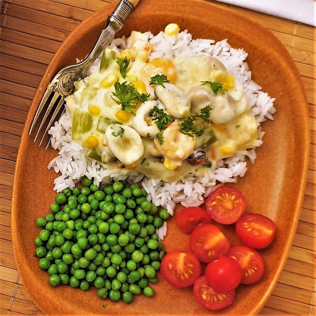 Overhead shot of a plate of seafood a la king, on a plate of rice, with peas and tomatoes.