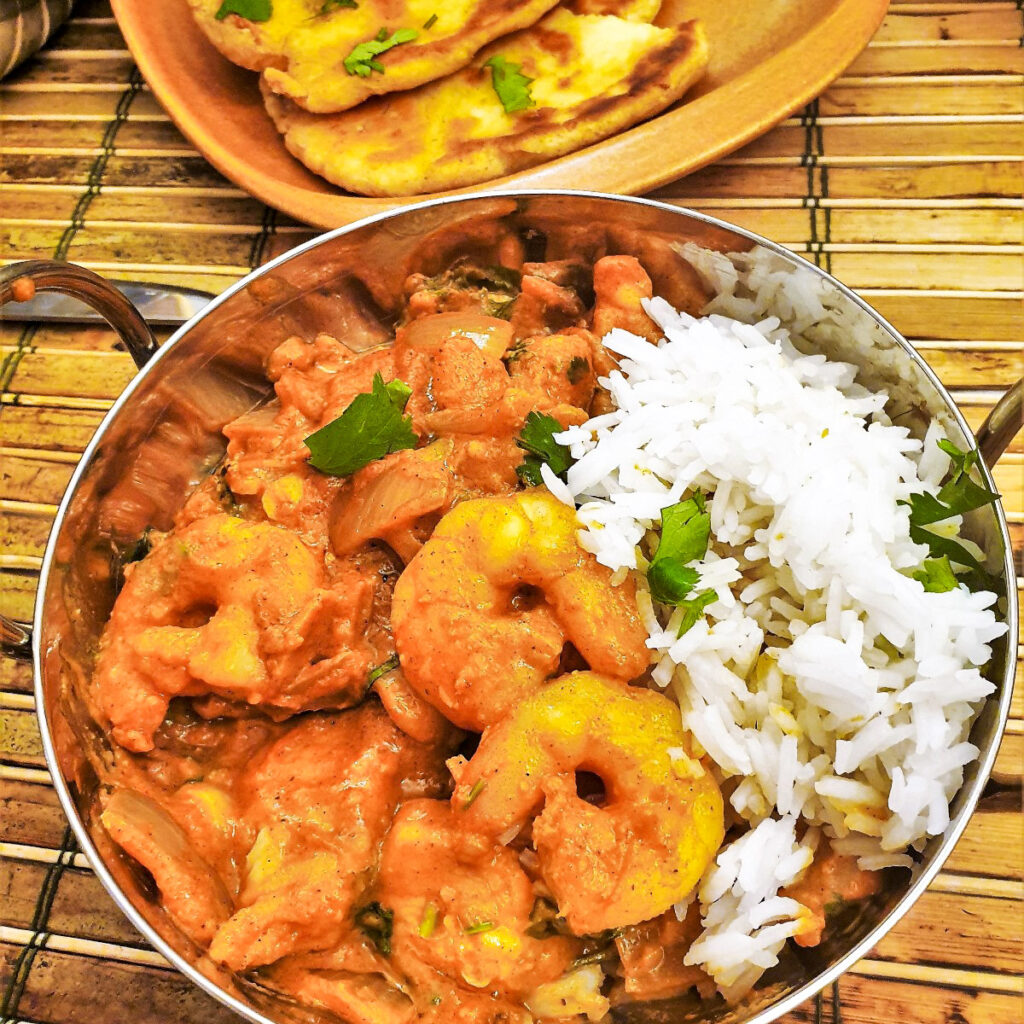 Kashmiri chicken and prawn curry in a dish in front of a plate of homemade buttered naan bread.