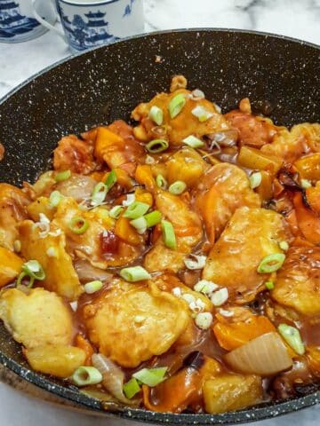 Overhead shot of a pan of sweet and sour chicken pieces garnished with spring onions in a thick sticky sauce.