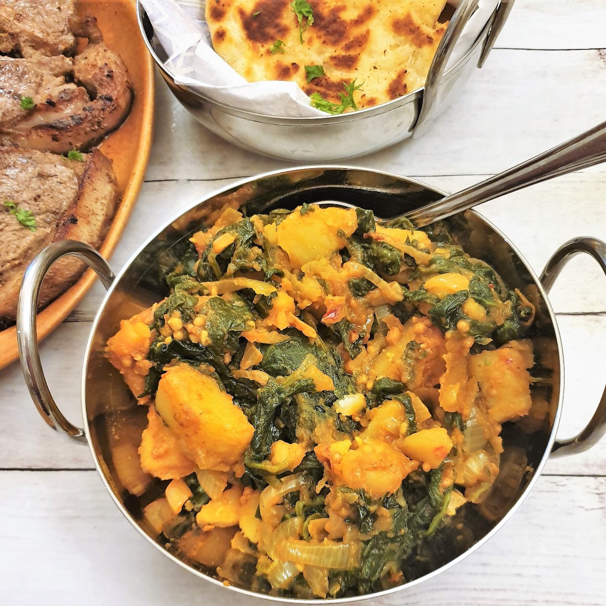 A dish of saag aloo next to tandoori lamb chops and naan bread.