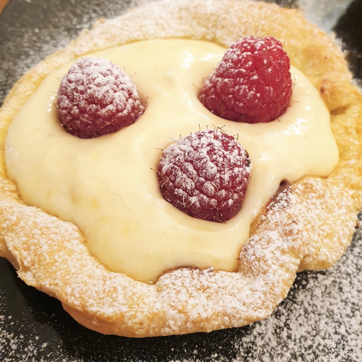 A raspberry custard tart with 3 raspberries, sprinkled with icing sugar.