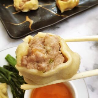 A steamed pork and shrimp dumpling held over a bowl of dipping sauce with a pair of chopsticks.