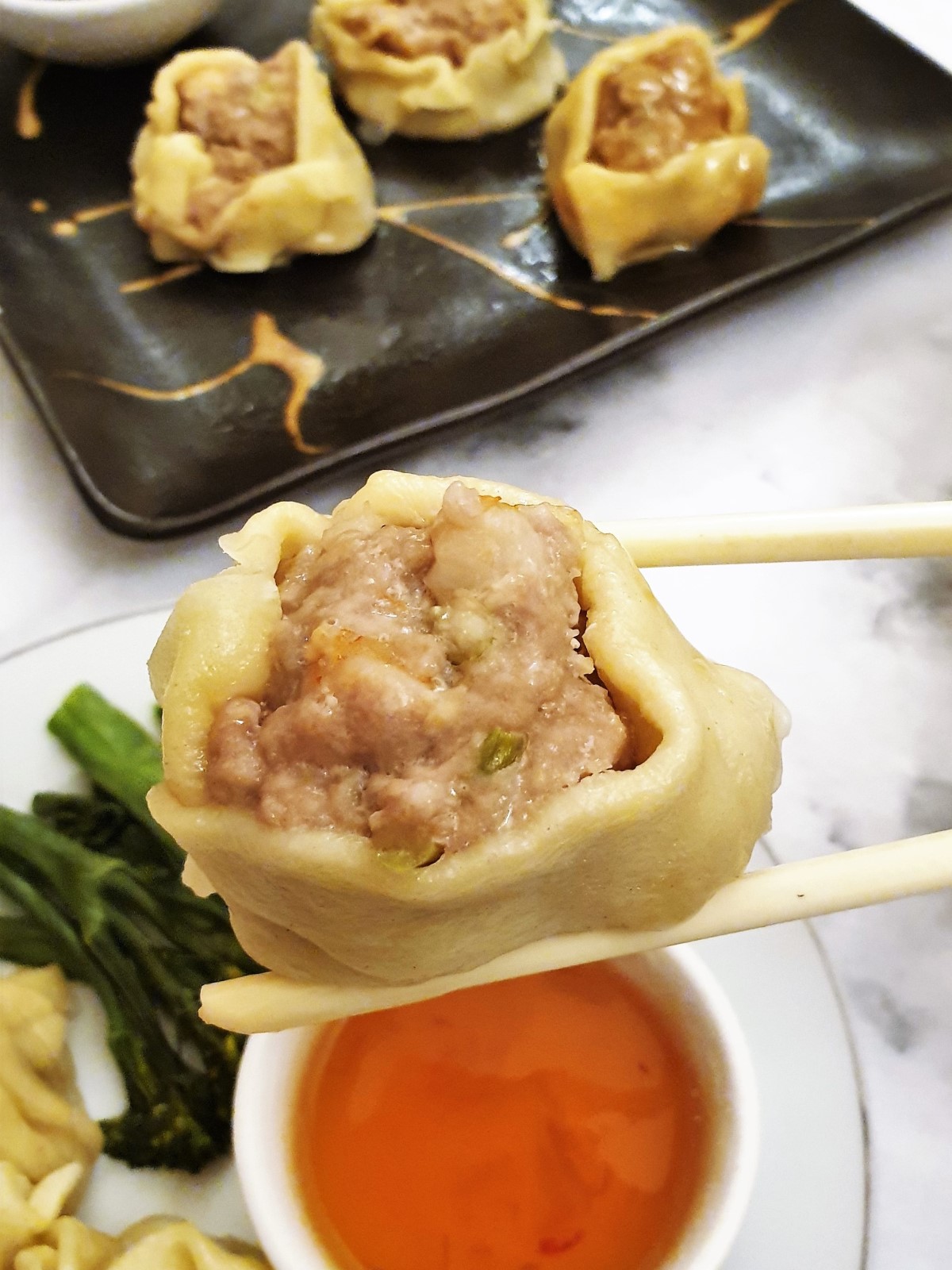 A steamed pork and shrimp dumpling held over a bowl of dipping sauce with a pair of chopsticks.
