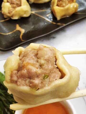 A steamed pork and shrimp dumpling held over a bowl of dipping sauce with a pair of chopsticks.