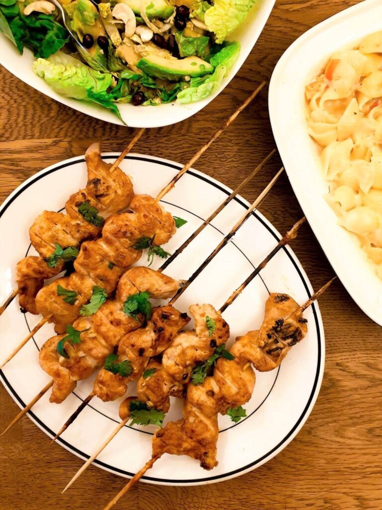 Batbequed chicken satay skewers on a plate next to bowls of salad..
