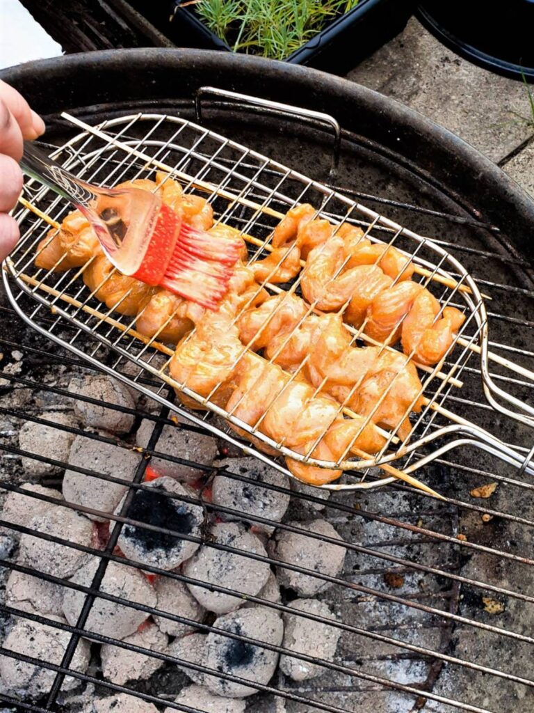 Satay chicken skewers being brushed with marinade on a barbeque grill.