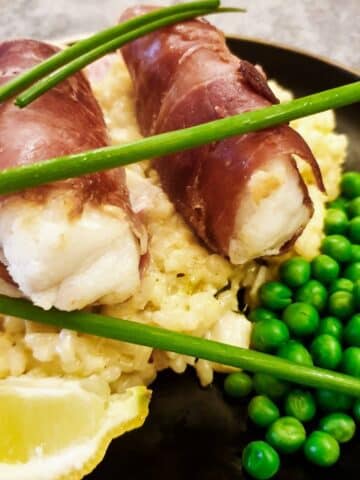 Closeup of two monkfish fillets wrapped in serrano ham on a bed of risotto with peas.