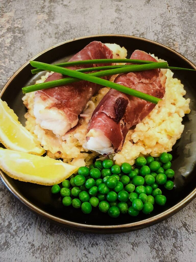 Two pieces of serrano-wrapped monkfish on a bed of lobster risotto with peas and two slices of lemon.