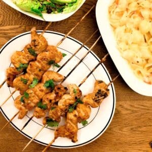 Barbequed chicken satay skewers on a plate next to bowls of salad..