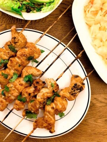 Barbequed chicken satay skewers on a plate next to bowls of salad..