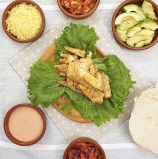 A pile of spicy fish on a plate of lettuce, surrounded by bowls of taco toppings and soft flour tortillas.