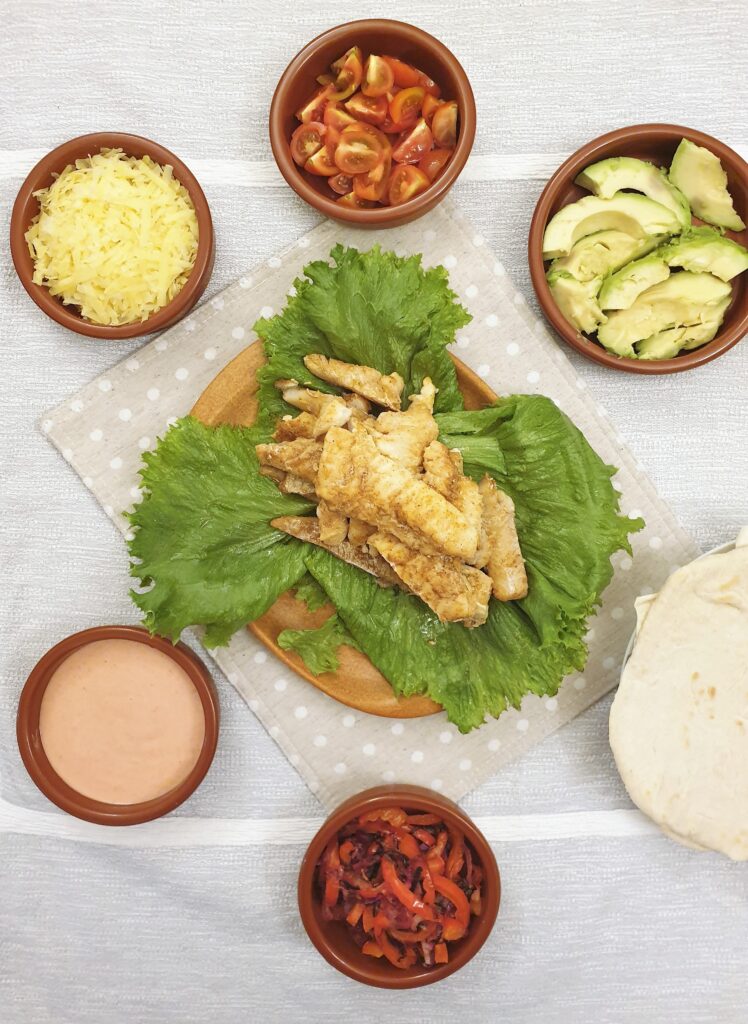 A pile of spicy fish on a plate of lettuce, surrounded by bowls of taco toppings and soft flour tortillas.