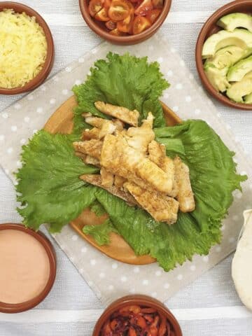 A pile of spicy fish on a plate of lettuce, surrounded by 6 small bowls of taco toppings and soft flour tortillas.