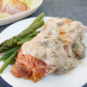 A bacon-wrapped chicken breast on a white plate, smothered in mushroom sauce with a side of asparagus spears.