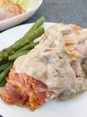 A bacon-wrapped chicken breast on a white plate, smothered in mushroom sauce with a side of asparagus spears.
